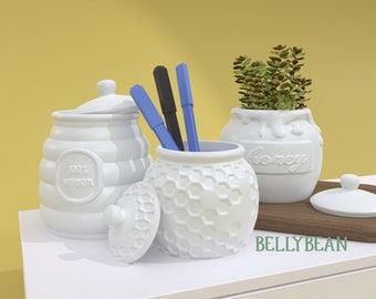 May include: White ceramic honey pot with a honeycomb design and the word "honey" on the front. A white ceramic jar with a honeycomb design and the text "100% NATURAL" on the front. A white ceramic jar with a honeycomb design and a lid. Three blue and black pens are in the jar. A small green plant is in the honey pot.