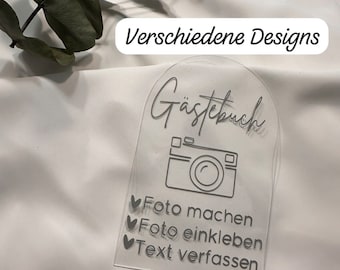 Pode incluir: Uma placa de acrílico transparente com o texto "Gästebuch" e um ícone de câmera. A placa também inclui instruções para os convidados tirarem uma foto, colarem-na e escreverem uma mensagem. O texto "Verschiedene Designs" é visível no fundo.