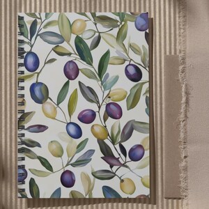 May include: Spiral-bound notebook featuring a watercolor olive branch design with green leaves and purple, blue, and yellow olives. The notebook rests on a textured, beige surface, with a shadow cast across the scene.