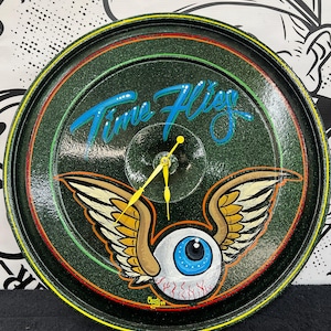 May include: A circular, green clock with the words "Time Flies" in blue script. The clock face features a winged eyeball design with a blue iris and yellow clock hands. The clock has a black, yellow, and red border.