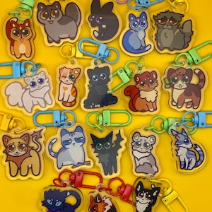 May include: A collection of cat-themed keychains with various designs and colors. Each keychain features a different cartoon cat illustration, attached to a colored clasp. The keychains are arranged on a bright yellow background.