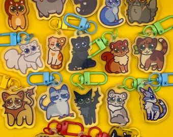 May include: A collection of cat-themed keychains with various designs and colors. Each keychain features a different cartoon cat illustration, attached to a colored clasp. The keychains are arranged on a bright yellow background.