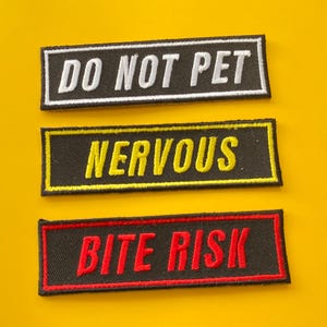 Może przedstawiać: Trzy prostokątne naszywki z haftowanym tekstem na żółtym tle. Górna naszywka głosi "DO NOT PET" w kolorze białym na czarnym tle. Środkowa naszywka głosi "NERVOUS" w kolorze żółtym na czarnym tle. Dolna naszywka głosi "BITE RISK" w kolorze czerwonym na czarnym tle.