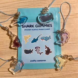 Op de afbeelding: Een lichtblauw zakje met het label SHARK GUMMIES MYSTERY ACRYLIC PHONE CHARM van crafty cameron, omringd door zes doorzichtige acryl haaienbedels in verschillende kleuren en soorten, elk bevestigd aan een lichtblauw koord met een zilveren sluiting.