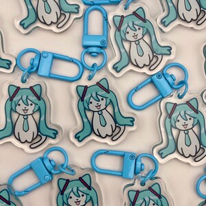 May include: Clear acrylic keychains featuring a cartoon character with long, turquoise hair and a matching tie. Each keychain has a light blue clasp. The character has a smiling expression and is surrounded by other keychains.