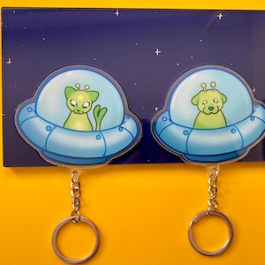 Puede incluir: Dos llaveros con extraterrestres de dibujos animados en ovnis azules. Cada llavero tiene una cadena plateada y un anillo plateado redondo. Los extraterrestres son verdes y tienen ojos grandes. El fondo es azul oscuro con estrellas blancas.