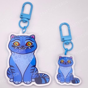 Peut inclure: Deux porte-clés en acrylique représentant un chat bleu avec des yeux jaunes, des rayures bleu foncé et des crocs visibles. Chaque porte-clés comprend un mousqueton en plastique bleu clair et un anneau, avec le texte CraftyCameron visible.