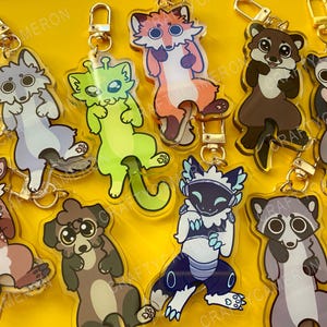 Op de afbeelding: Een verzameling van acht acryl sleutelhangers met cartoon dierfiguren waaronder een wolf, alien kat, vos, otter, opossum, hert, hond en wasbeer, elk bevestigd aan een goudkleurige metalen karabijnhaak en draaibare sluiting.
