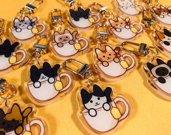 Puede incluir: Llaveros acrílicos de gatos en varios colores, cada uno dentro de una taza de té blanca con una bolsita de té amarilla. Los llaveros tienen cierres y anillos dorados. El fondo es amarillo.