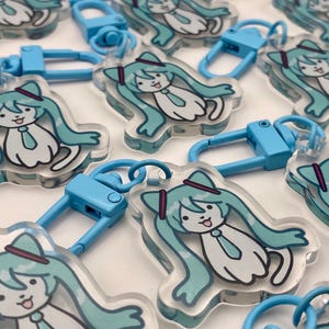 May include: A collection of clear acrylic keychains featuring a cartoon character with long, turquoise hair, a tie, and a hat. Each keychain has a blue clasp for attachment.