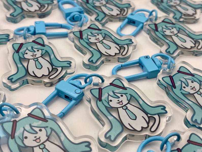 May include: A collection of clear acrylic keychains featuring a cartoon character with long, turquoise hair, a tie, and a hat. Each keychain has a blue clasp for attachment.