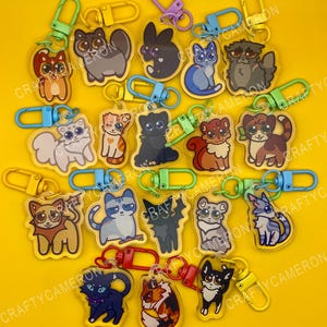 Pode incluir: Uma coleção de porta-chaves de acrílico com vários gatos ilustrados de diversas cores e padrões de pelagem, cada um preso a um fecho mosquetão de plástico colorido. O texto CRAFTYCAMERON é visível num padrão repetido.