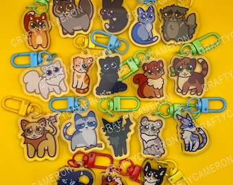 Puede incluir: Una colección de llaveros de acrílico con varios gatos ilustrados de diversos colores y patrones de pelaje, cada uno unido a un cierre de mosquetón de plástico de colores. El texto CRAFTYCAMERON es visible en un patrón repetido.