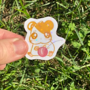 以下が含まれることがあります： ピンクのサクランボを持った、漫画のハムスターの白と金のキラキラステッカー。ハムスターは大きな目と茶色の鼻をしています。