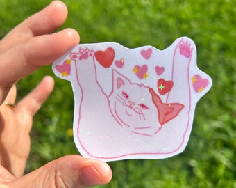 Puede incluir: Una pegatina rosa y blanca de un gato con las patas en el aire, rodeado de corazones rosas y purpurina. El gato tiene una expresión de sorpresa en su rostro.