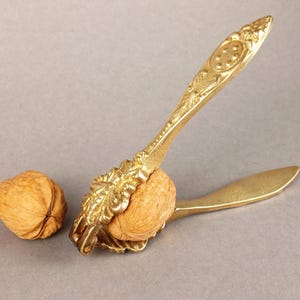 May include: A gold-coloured nutcracker with intricate floral detailing is shown, gripping a walnut. The nutcracker is open, ready to crack the nut. Another walnut sits to the left. The background is a solid grey.