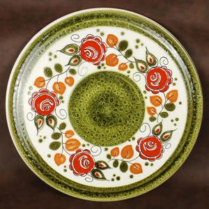 Könnte beinhalten: Runder Keramikteller mit weißem Hintergrund, grünem Rand und einem grünen Mittelkreis. Verziert mit roten Rosen, orangefarbenen Blättern und grünem Laub, das ein Blumenmuster bildet. Der Teller hat eine Vintage-Ästhetik.