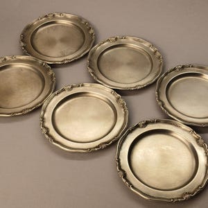 Puede incluir: Seis platos antiguos de color plateado con bordes festoneados ornamentados. Los platos est&aacute;n dispuestos en un patr&oacute;n ligeramente desplazado, mostrando sus bordes decorativos y superficies reflectantes. Un conjunto cl&aacute;sico para cenas formales.