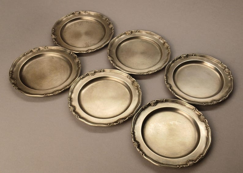 Puede incluir: Seis platos antiguos de color plateado con bordes festoneados ornamentados. Los platos est&aacute;n dispuestos en un patr&oacute;n ligeramente desplazado, mostrando sus bordes decorativos y superficies reflectantes. Un conjunto cl&aacute;sico para cenas formales.