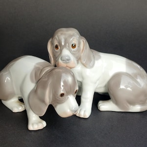 Peut inclure: Deux figurines de chiens en céramique. L'une est assise, l'autre est accroupie. Les deux ont des marques grises et blanches, avec des yeux et des nez bruns. Les figurines sont sur une surface sombre.