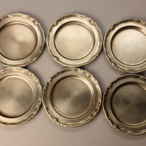 Puede incluir: Seis platos de color plateado con bordes festoneados ornamentados. Los platos tienen un dise&ntilde;o circular simple en el centro. Los platos est&aacute;n dispuestos en dos filas de tres sobre un fondo neutro. Los platos son probablemente para servir comida.