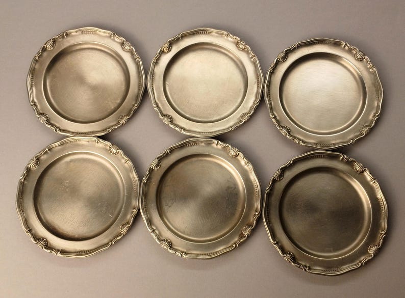 Puede incluir: Seis platos de color plateado con bordes festoneados ornamentados. Los platos tienen un dise&ntilde;o circular simple en el centro. Los platos est&aacute;n dispuestos en dos filas de tres sobre un fondo neutro. Los platos son probablemente para servir comida.