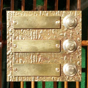 Può includere: Una placca per campanello in ottone d'epoca con tre targhette rettangolari e tre pulsanti rotondi. La placca ha una superficie strutturata ed è montata con viti. L'ottone presenta una calda patina invecchiata.