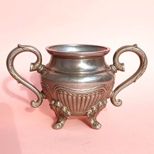 Pode incluir: Açucareiro antigo prateado com duas alças ornamentadas e três pés decorativos. O açucareiro apresenta um design em relevo detalhado ao redor do corpo e uma borda lisa. Uma peça clássica para servir açúcar ou outros itens pequenos.