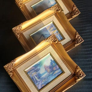 Op de afbeelding: Drie ingelijste landschapsschilderijen met sierlijke gouden lijsten. Elk schilderij toont een serene buitenscène met bomen, water en een blauwe lucht. De lijsten hebben decoratieve hoekaccenten.