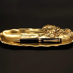 May include: A gold-toned metal tray with an elongated, wavy-edged shape featuring a raised floral relief of daisies and leaves on one side. A black ballpoint pen with gold-colored accents rests horizontally in the center of the tray.