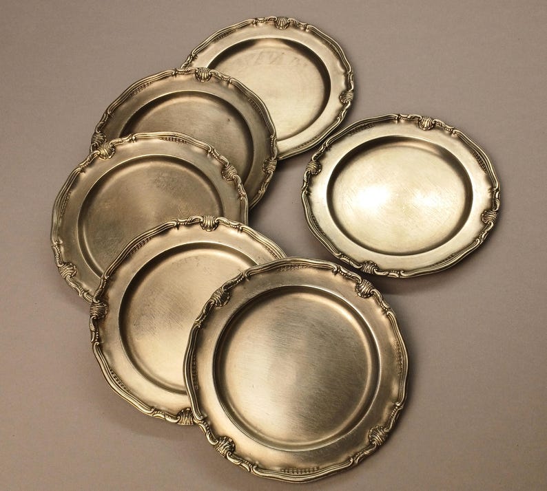 Puede incluir: Un conjunto de platos antiguos de color plateado con bordes festoneados ornamentados. Los platos est&aacute;n dispuestos en un patr&oacute;n ligeramente superpuesto, mostrando sus bordes decorativos y superficies reflectantes. Los platos son probablemente para servir comida.