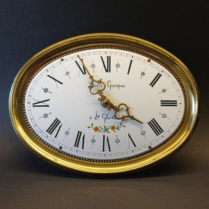 May include: An oval wall clock with a polished gold-tone frame and a white face. The dial features black Roman numerals, ornate gold-tone hands, and the text Belle Epoque à St Ghislain. A small floral motif with orange flowers sits at the bottom.