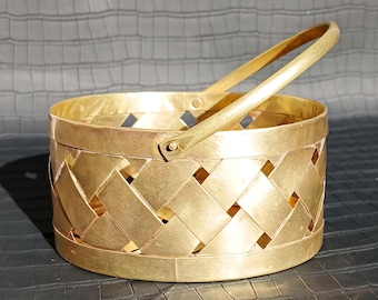May include: A gold-colored basket with a woven design and a handle. The basket has a round shape and is made of metal. The handle is attached to the top of the basket with rivets.