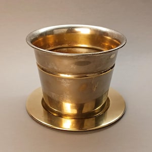 Puede incluir: Una taza de metal plateado y dorado con su platillo. La taza tiene una parte superior ancha y un cuerpo cilíndrico. El platillo es redondo y plano. El metal tiene un acabado pulido que refleja la luz.