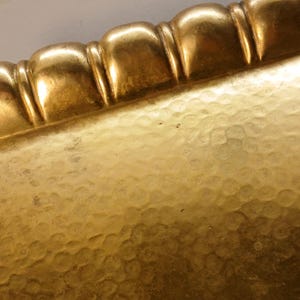 May include: Close-up of a gold-colored rectangular tray with a textured surface and a decorative, scalloped edge. The tray has a hammered appearance, reflecting light and creating a warm, antique aesthetic.