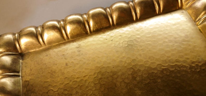 May include: Close-up of a gold-colored rectangular tray with a textured surface and a decorative, scalloped edge. The tray has a hammered appearance, reflecting light and creating a warm, antique aesthetic.