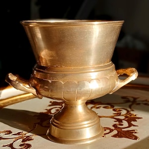 May include: A gold-coloured metal chalice with a wide, flared top and a rounded base. The chalice has two ornate handles and sits on a pedestal. The surface has a slightly aged appearance.