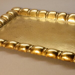 May include: A rectangular gold-colored tray with a hammered surface. The tray's border is adorned with a repeating, rounded design. The tray is likely used for serving or display purposes.