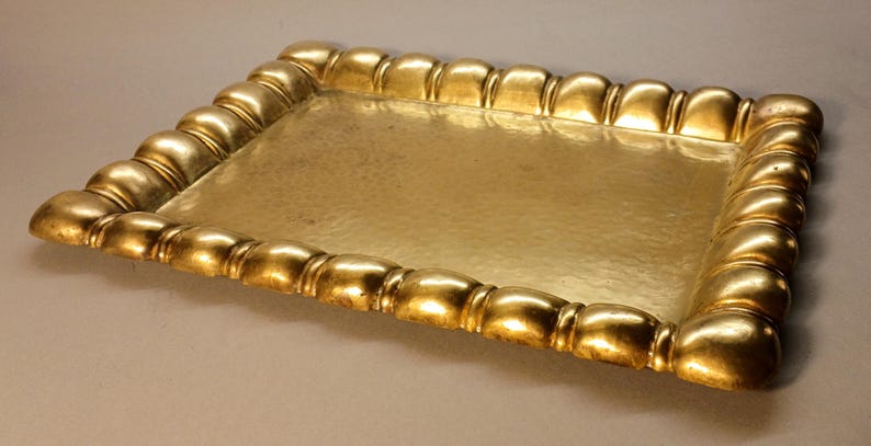 May include: A rectangular gold-colored tray with a hammered surface. The tray's border is adorned with a repeating, rounded design. The tray is likely used for serving or display purposes.