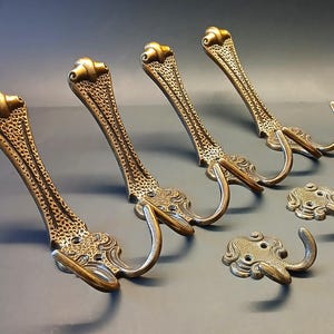 Op de afbeelding: Een set van zes messingkleurige metalen haken, bestaande uit vier lange, slanke wandhaken met een getextureerd oppervlak en krulvormige bovenkanten, samen met twee kleinere, compacte dubbele haken met bijpassende details en zichtbare schroefgaten.