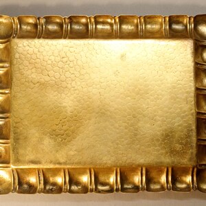 May include: A rectangular, gold-colored tray with a textured surface and a decorative border. The border features a series of rounded, square-like shapes. The tray's surface has a hammered or dimpled appearance, adding visual interest.