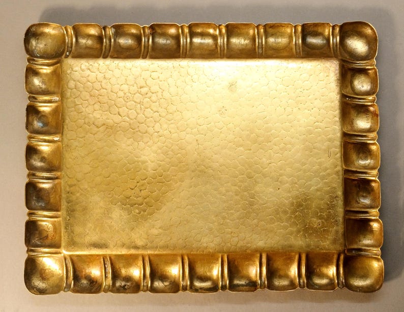 May include: A rectangular, gold-colored tray with a textured surface and a decorative border. The border features a series of rounded, square-like shapes. The tray's surface has a hammered or dimpled appearance, adding visual interest.