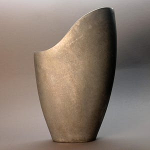May include: A tall, metallic vase with a unique, asymmetrical design. The vase has a smooth, textured surface and a wide opening on one side. The color is a muted silver-gray, giving it a modern and minimalist aesthetic.
