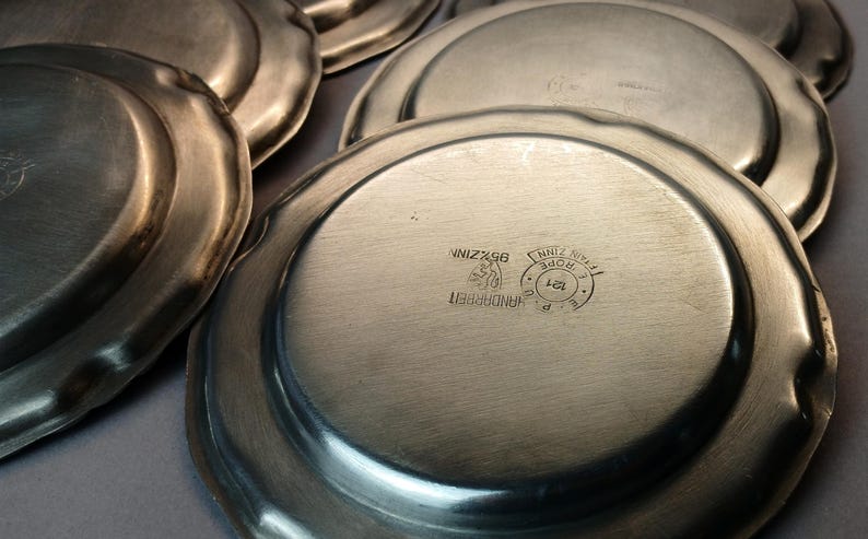 Puede incluir: Una colecci&oacute;n de platos antiguos de peltre con un borde ligeramente festoneado. Los platos tienen un acabado de metal cepillado y est&aacute;n estampados con marcas que incluyen "95% ZINN" y "HANDARBEIT".