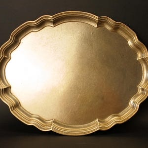 Puede incluir: Una bandeja de servicio metálica de forma ovalada con un acabado en tono dorado pulido. La bandeja presenta un borde elevado y festoneado con un diseño de reborde curvo continuo que crea un marco decorativo alrededor de la superficie central.