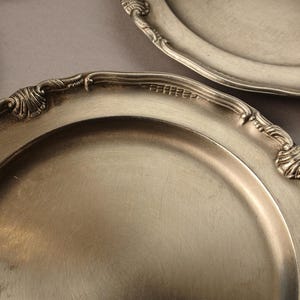 Puede incluir: Varias fuentes antiguas de color plateado con bordes ornamentados y en relieve se disponen sobre una superficie gris. Las fuentes tienen una superficie reflectante y un dise&ntilde;o cl&aacute;sico, lo que sugiere una cena formal o un uso decorativo.