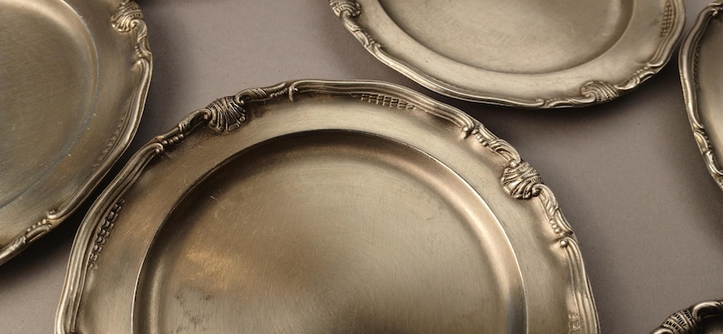 Puede incluir: Varias fuentes antiguas de color plateado con bordes ornamentados y en relieve se disponen sobre una superficie gris. Las fuentes tienen una superficie reflectante y un dise&ntilde;o cl&aacute;sico, lo que sugiere una cena formal o un uso decorativo.