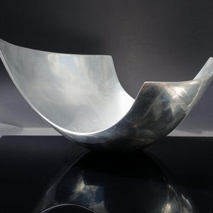 Op de afbeelding: Een gepolijste, zilverkleurige decoratieve kom met een gebogen, open ontwerp. De kom heeft een glad, reflecterend oppervlak en staat op een zwart oppervlak, waardoor een spiegeleffect ontstaat. De vorm van de kom doet denken aan een halve maan.