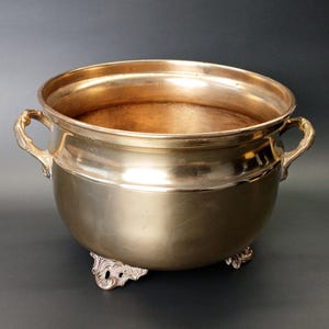 May include: A polished brass bowl with a smooth, rounded body, featuring two ornate, curved handles on opposite sides. The vessel rests on three decorative, scrolled feet and has a wide, open top with a smooth, reflective interior surface.