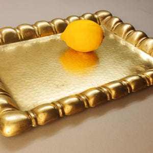 May include: A gold-colored metal tray with a hammered surface and a decorative, rounded border. A yellow lemon sits in the center of the tray, reflecting on the surface. The tray is rectangular and appears to be made of brass.